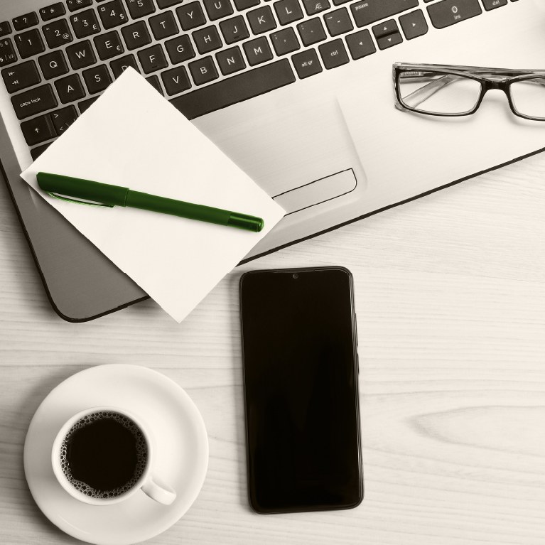 desk with laptop, phone notepad with green pen and a cup of coffee